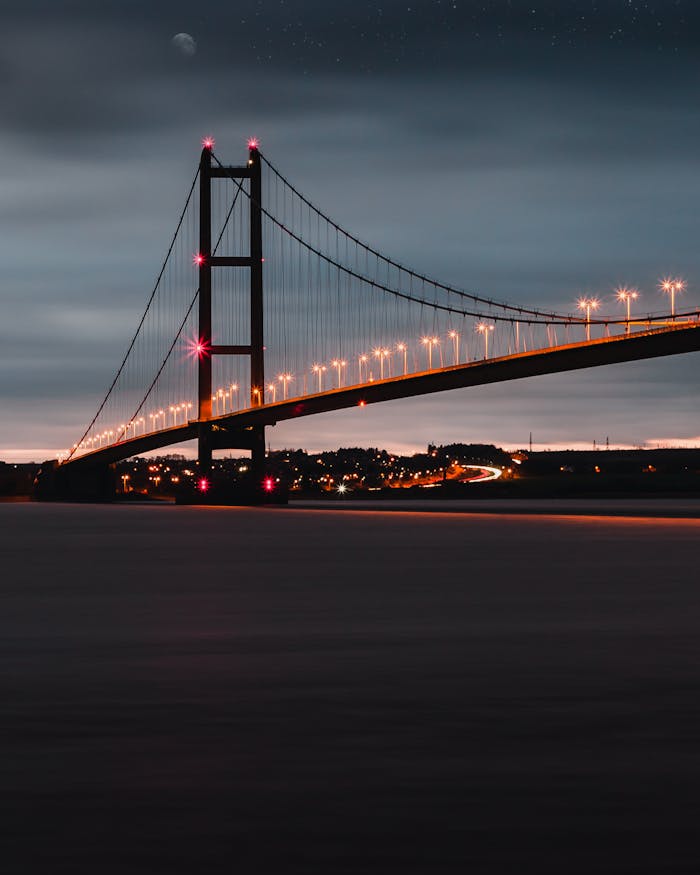 A dramatic night scene of a beautifully lit suspension bridge under a cloudy, starry sky.